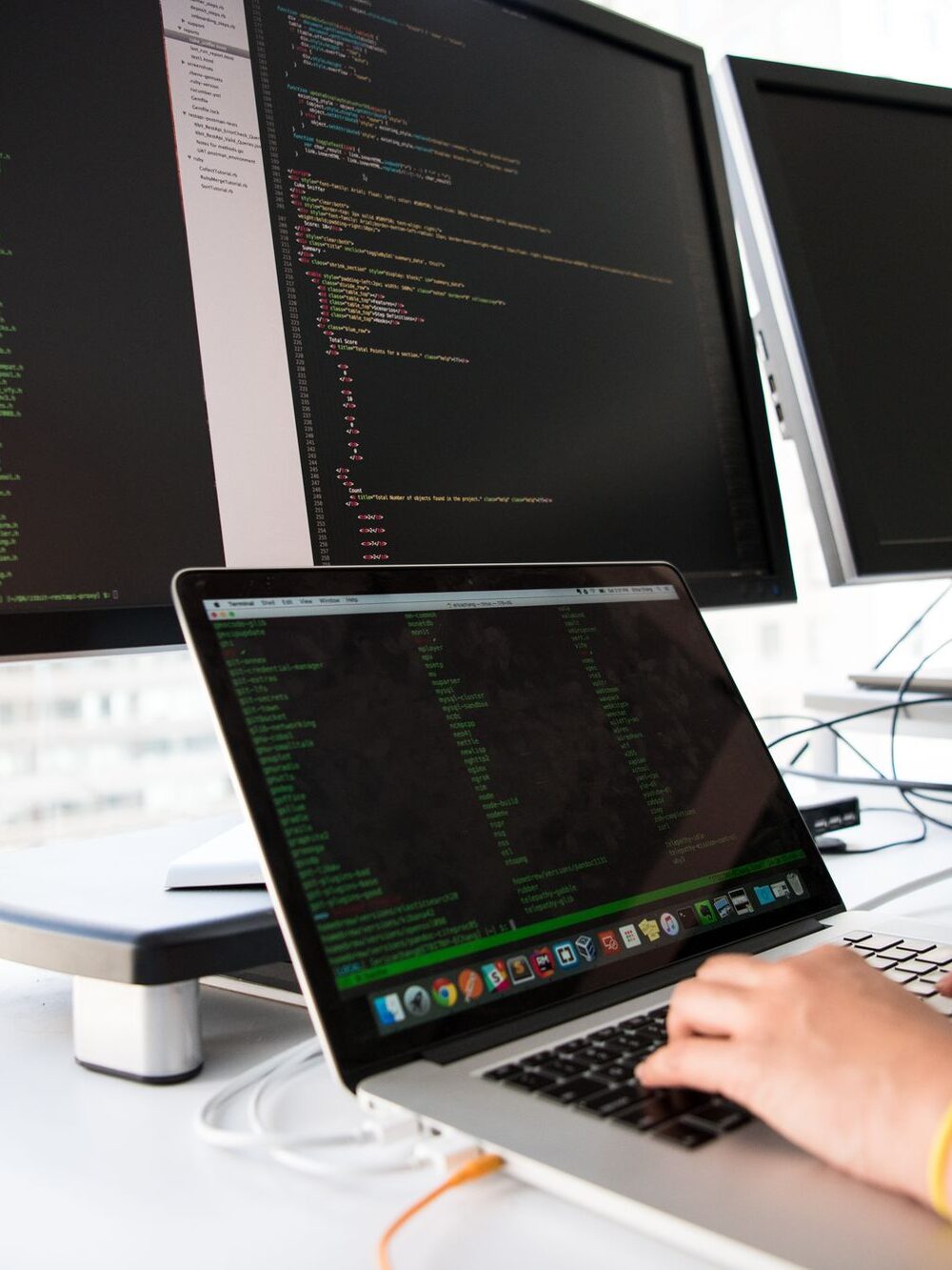 Mãos digitando em um notebook com código aberto em dois monitores.